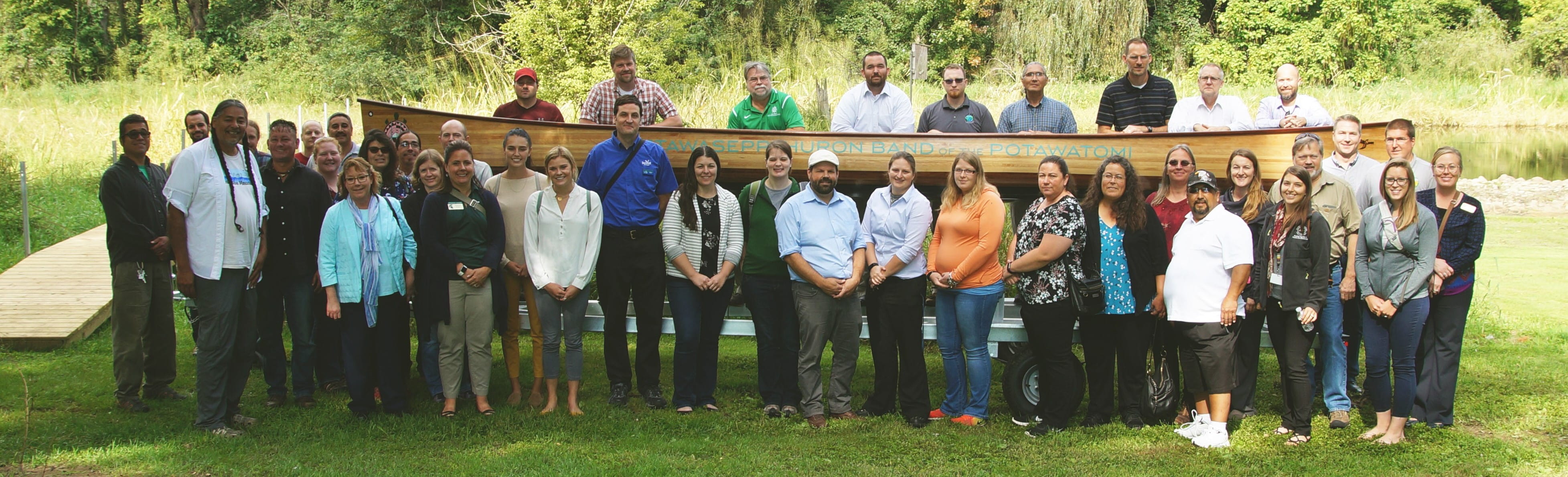 A group of about 50 people standing outside in front of a long wooden canoe