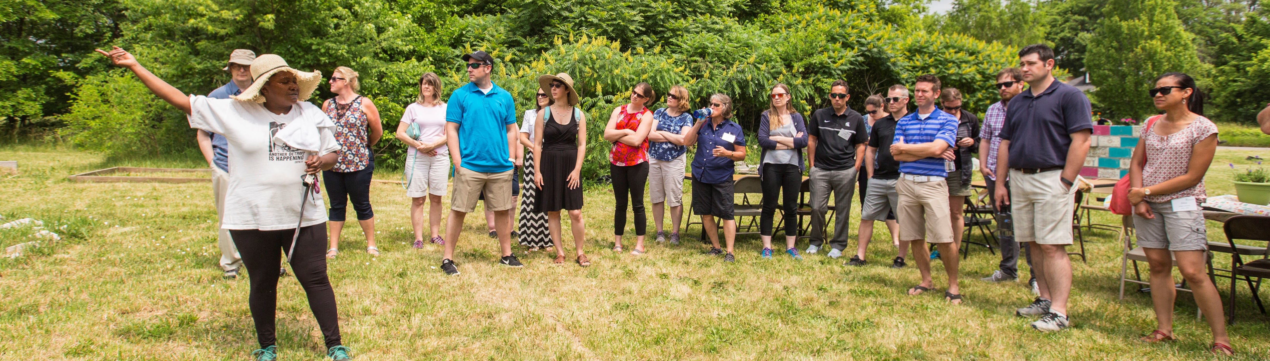 A woman gesturing to her left in a field with a crowd standing and listening