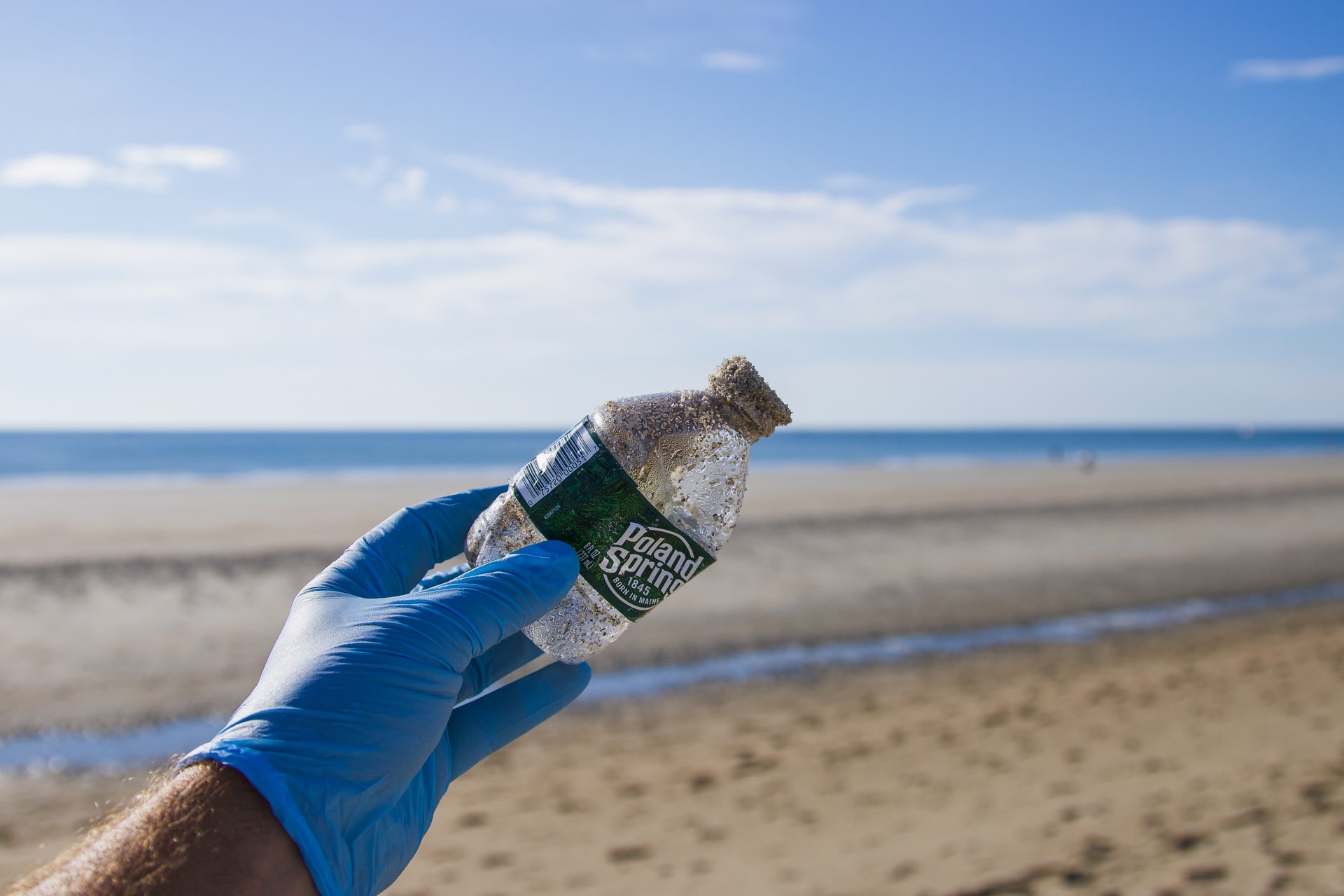 A hand wearing a rubber glove holding up a discarded plastic water bottle from a sandy beach