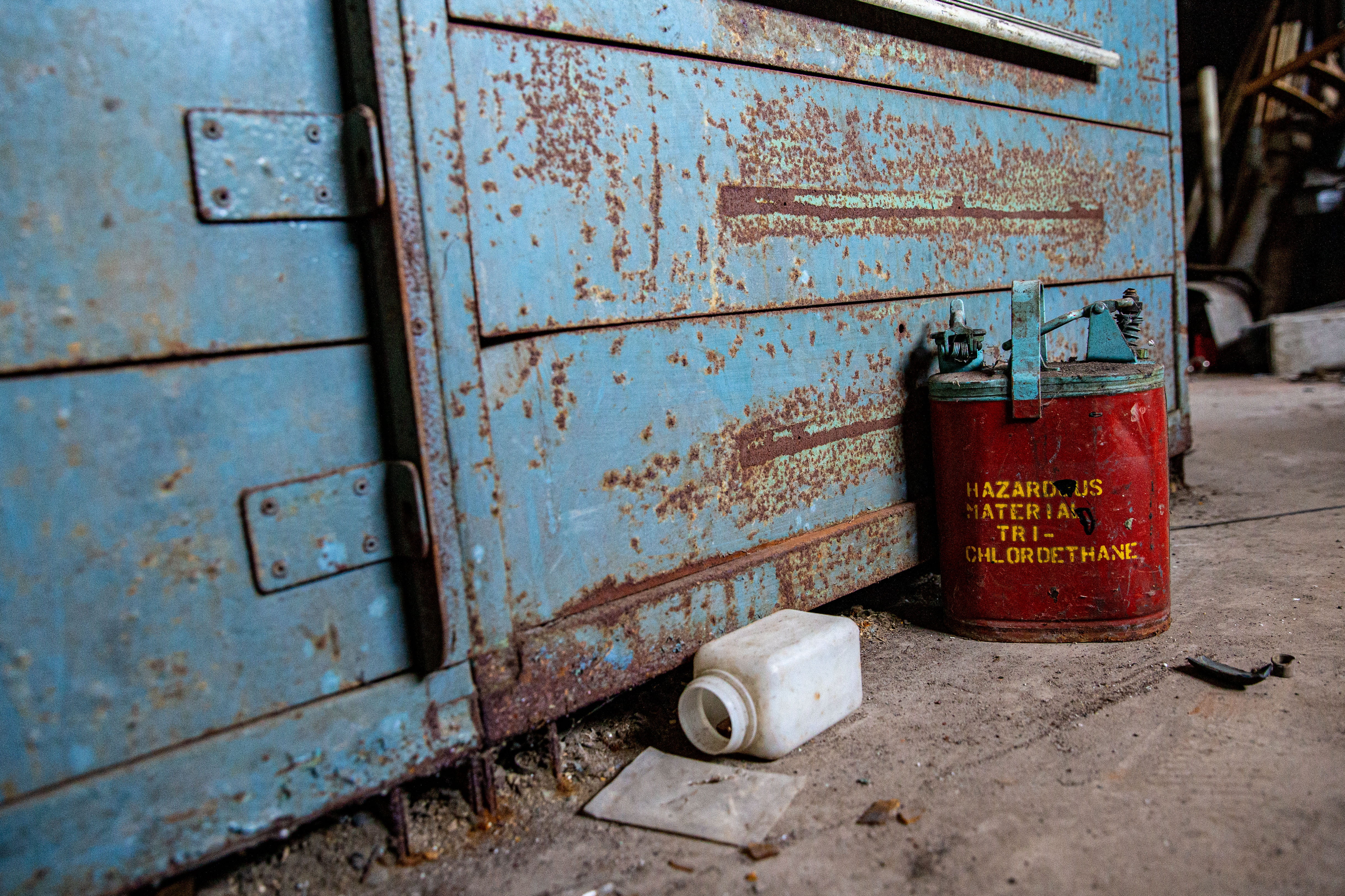 A red vintage metal jug reading Hazardous Material Tri-Chlordethane sits on a dirty and rusty warehouse floor