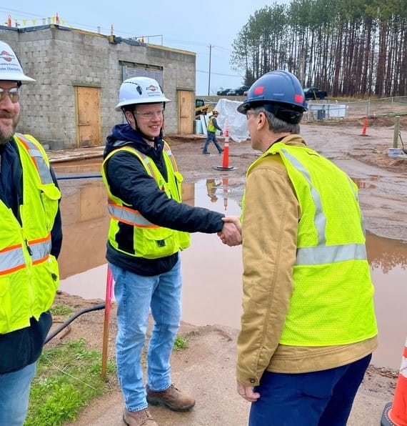 EGLE Director Phil Roos wearing a yellow construction vest and hardhat shaking the hand of a staff member also wearing a vest and hardhat, with construction on the WWTP in the background