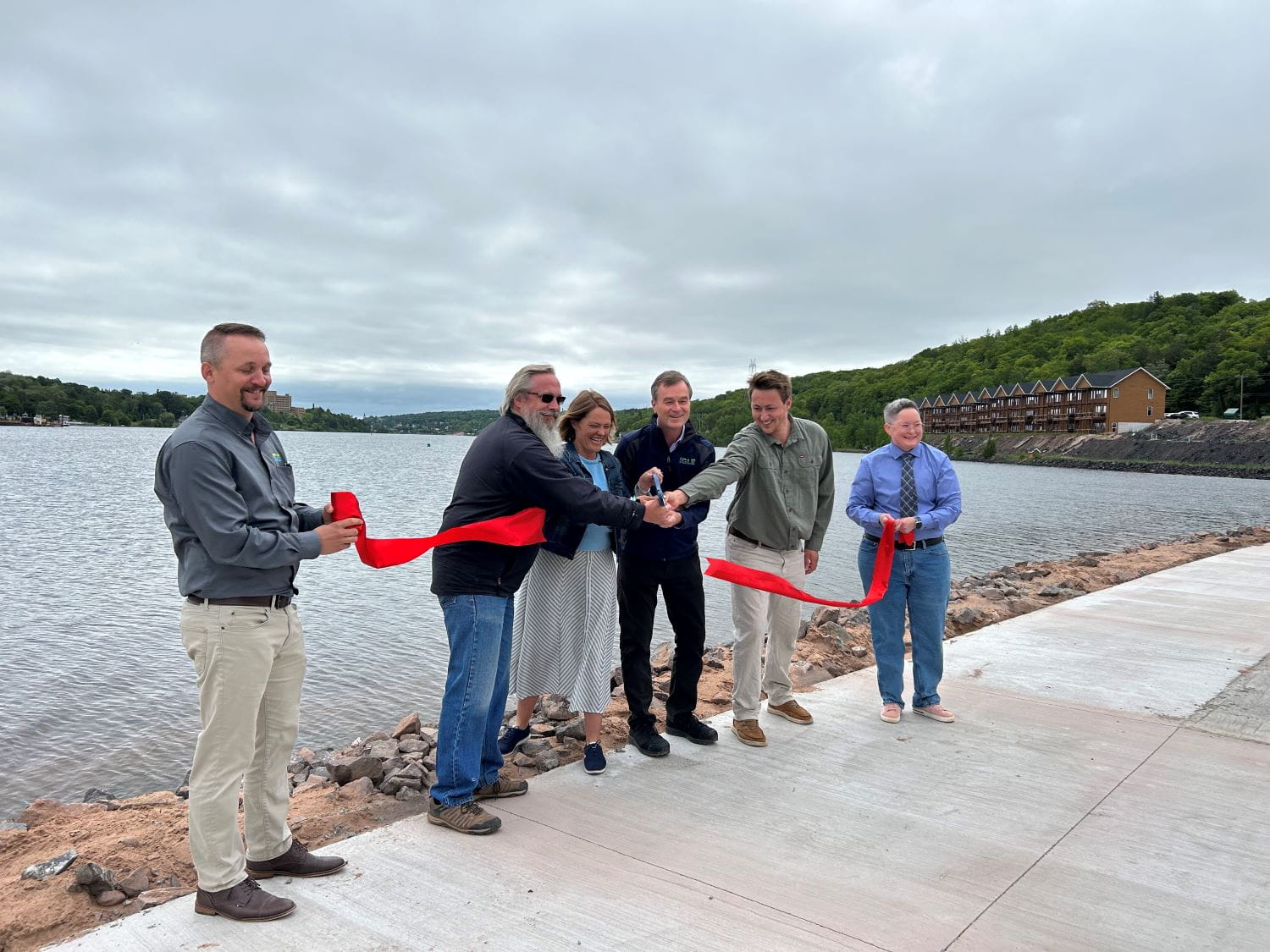 EGLE Director Phil Roos and 5 coworkers stand on the shore of Torch Lake holding a large pair of scissors cutting through a red ribbon