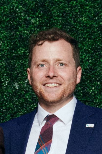 Cory Connolly, EGLE's Climate and Energy Advisor, in a navy blazer, white shirt, and red tie smiling against a greenery backdrop