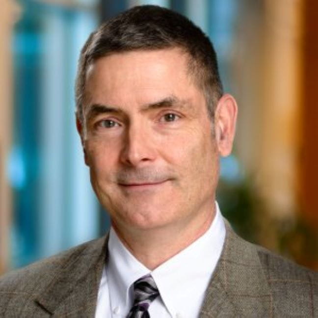 Hugh McDiarmid, Jr. wearing a white shirt, tie, and brown blazer and smiling