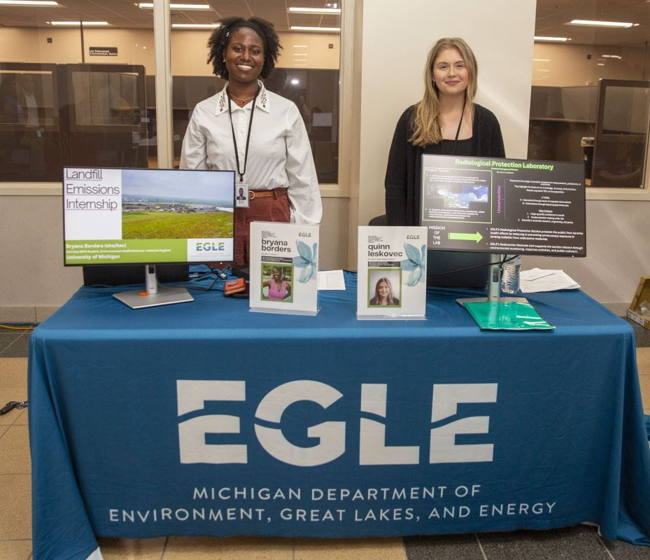 Bryana Borders and Quinn Leskovec are interns at a EGLE table, the table has computer screens showing protection lab information and land fill emissions internships.