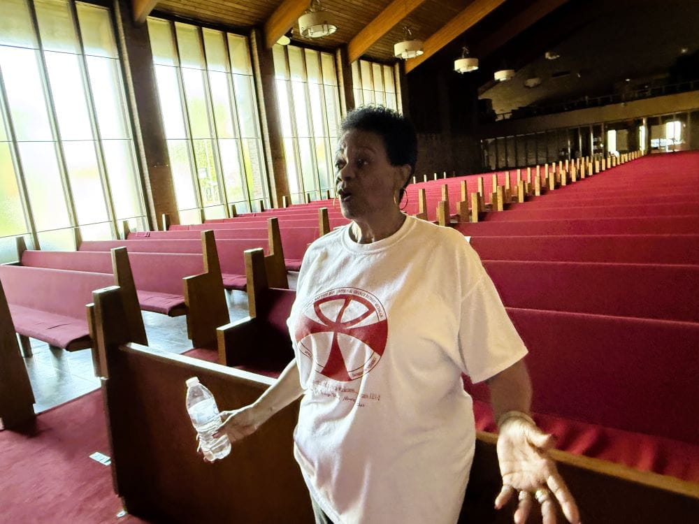 A woman standing in a church in front of rows of pews speaking emphatically