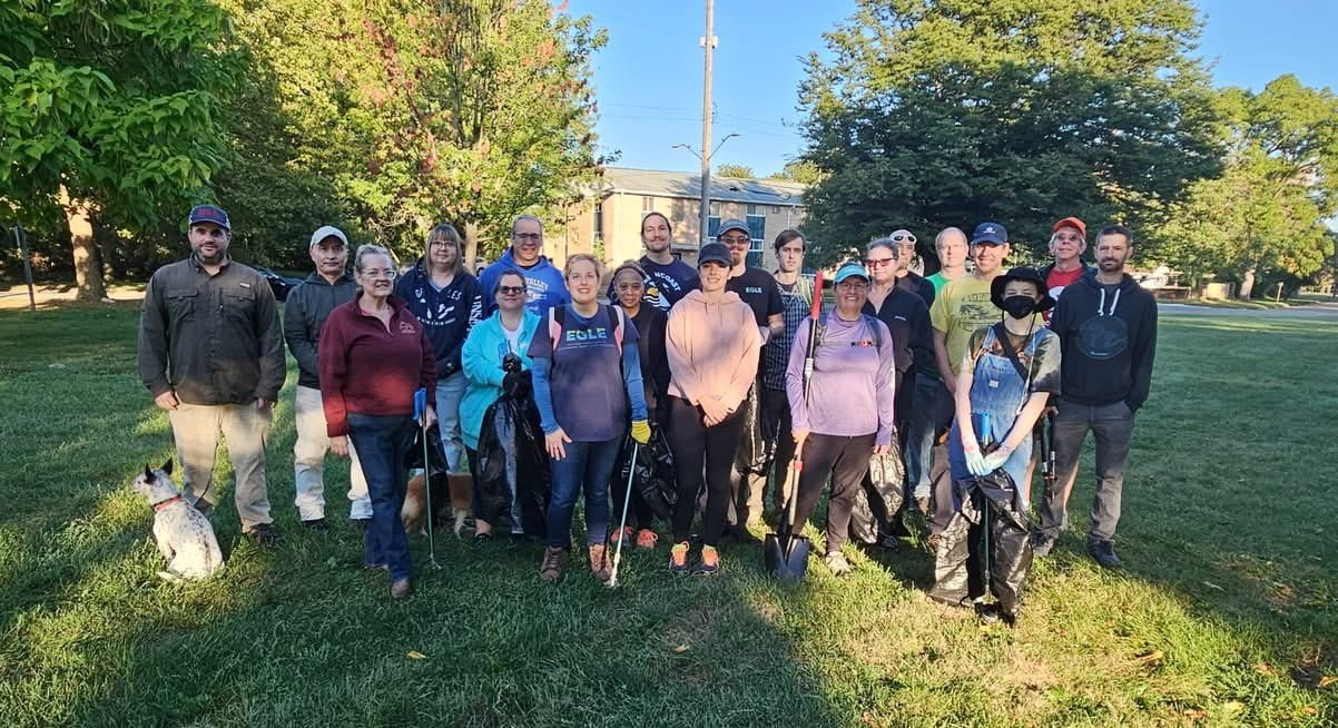 A group of about 25 volunteers pose standing on grass for a picture holding trash bags and garbage picks