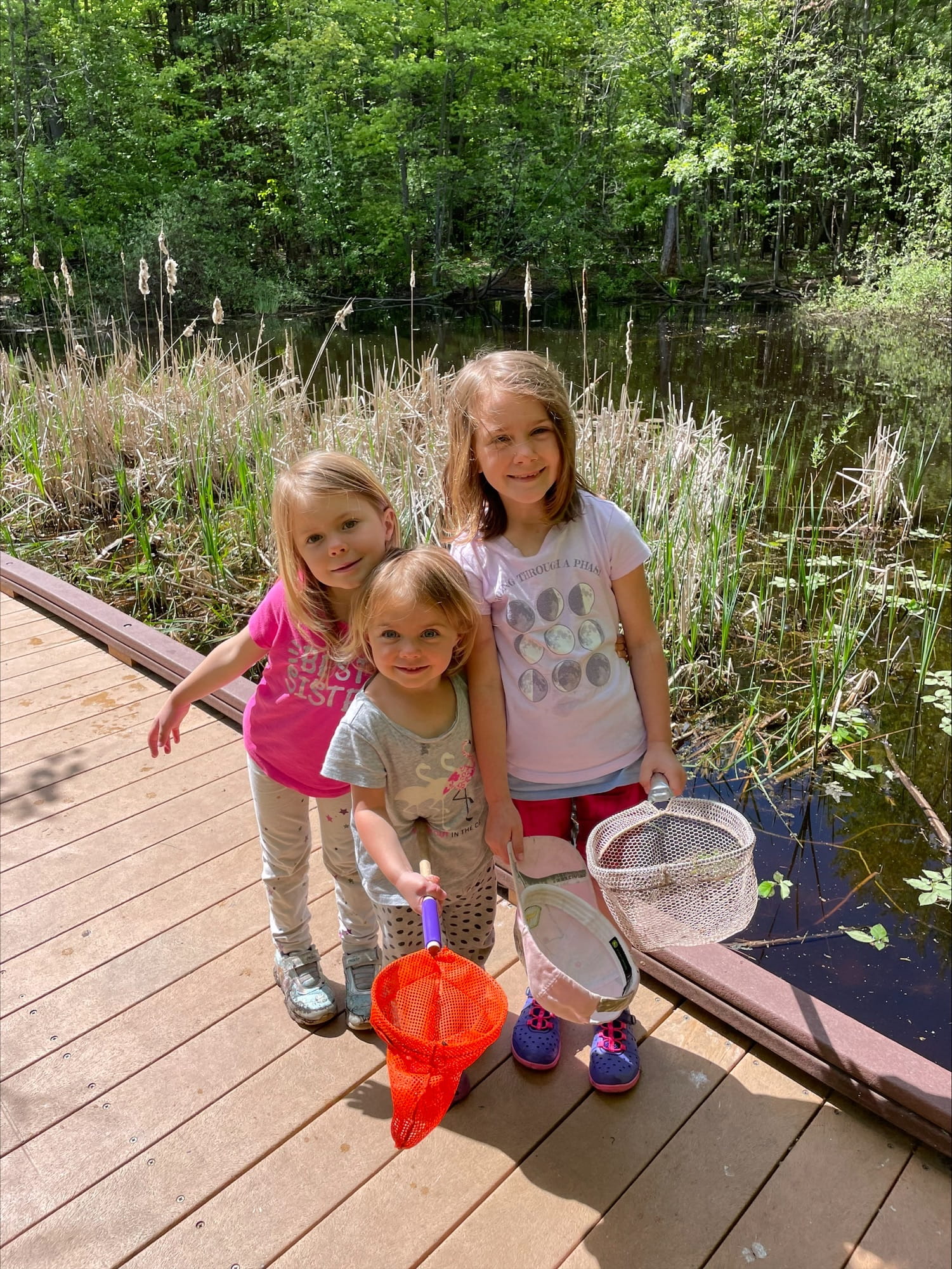 Autumn, Winter, and Audra Boothroyd, daughters of Jeremy Boothroyd, holding nets standing on a boardwalk over wetland. 