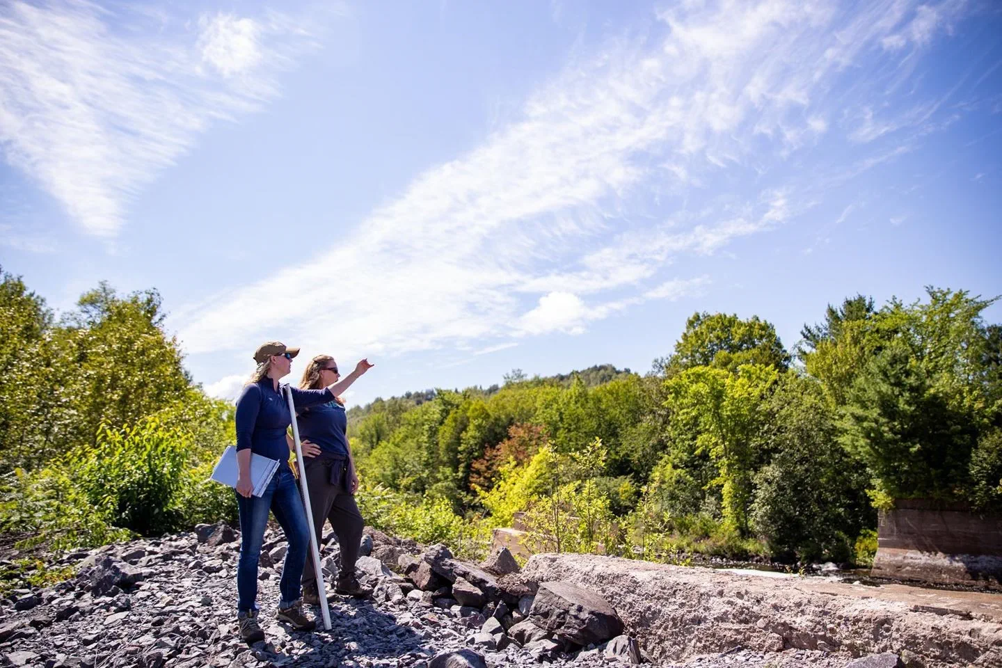 Two EGLE Water Resources staff members out doing field work on a rocky river bank pointing at the water in the distance on a nice sunny day
