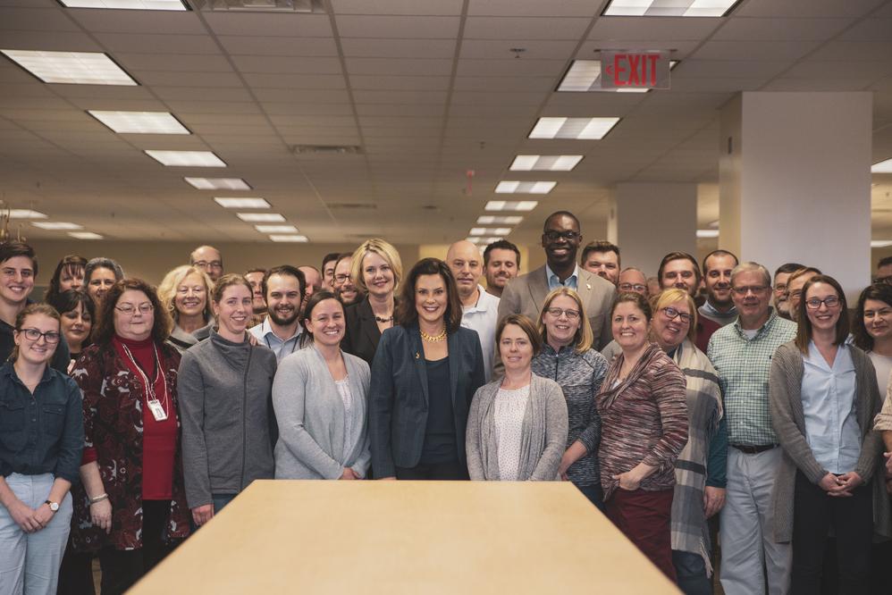 A large group of diverse EGLE staff stand around a table, smiling