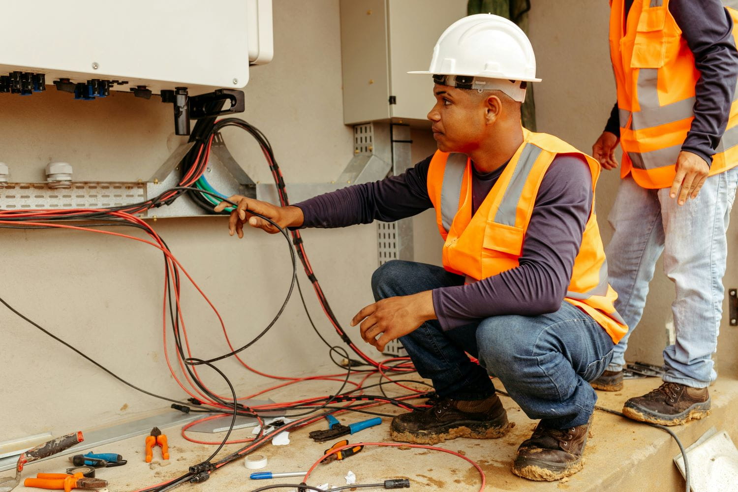 A renewable energy contractor wearing a hardhat and vest, squatting on the floor of a building, working on electrical wiring