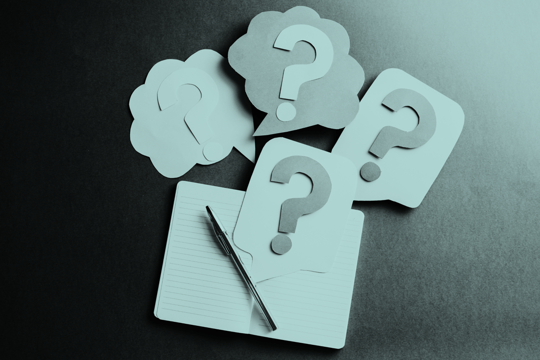Multiple paper cutouts of questions marks laid over a notebook and pen
