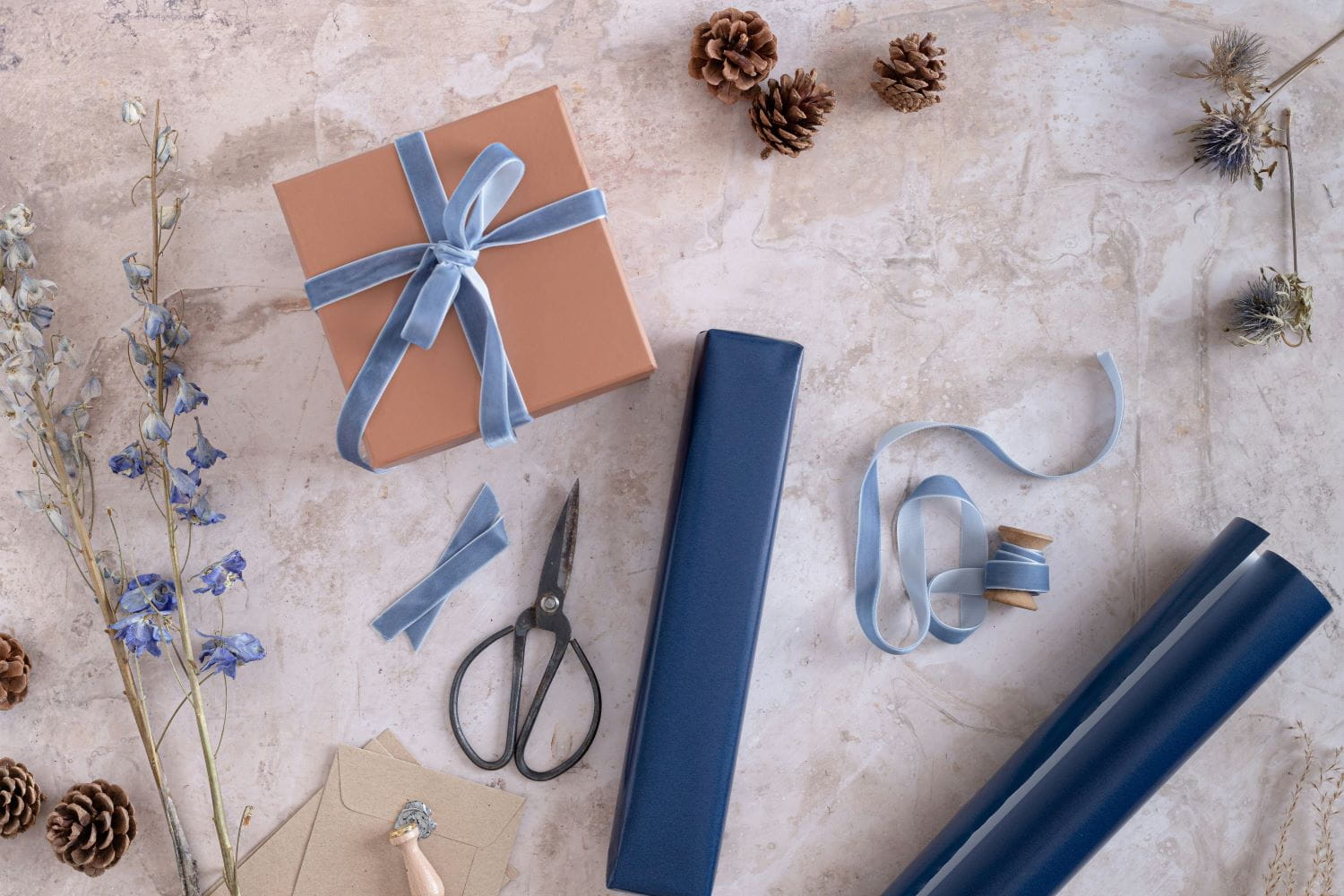 A box wrapped in brown packing paper with a cornflower blue velvet ribbon, with assorted giftwrapping supplies such as wrapping paper, scissors, boxes, etc. scattered across the table
