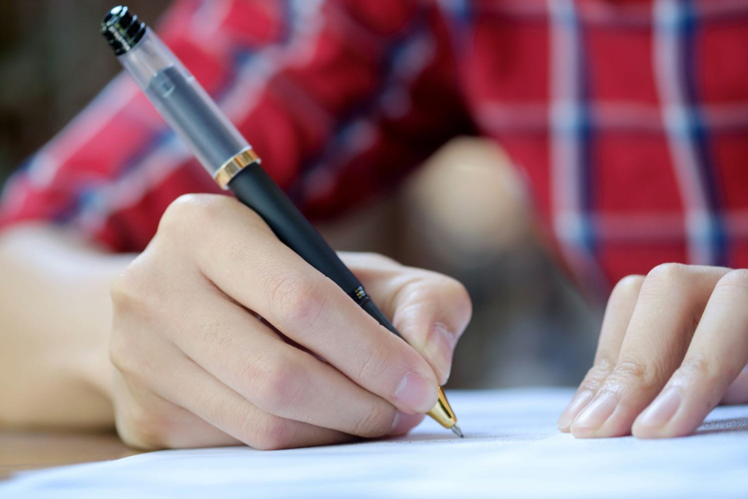 A person wearing a red plaid shirt signing a document on a desk