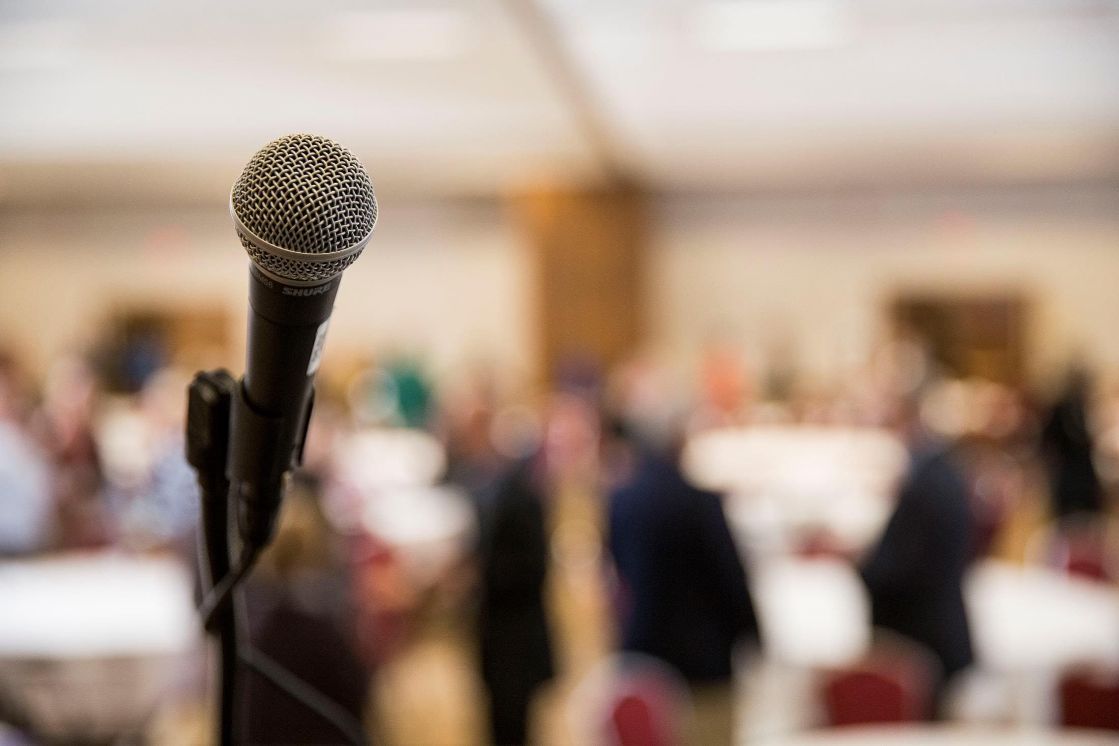 A single microphone in focus, with a blurred background of meeting attendees