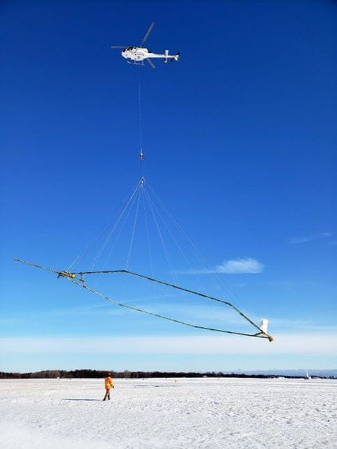 Helicopter carrying equipment in a blue sky while a person stands beneath this on the snow.
