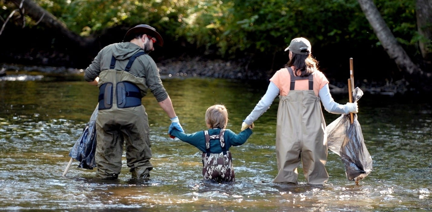 family wading through a river while carrying bags for cleanup