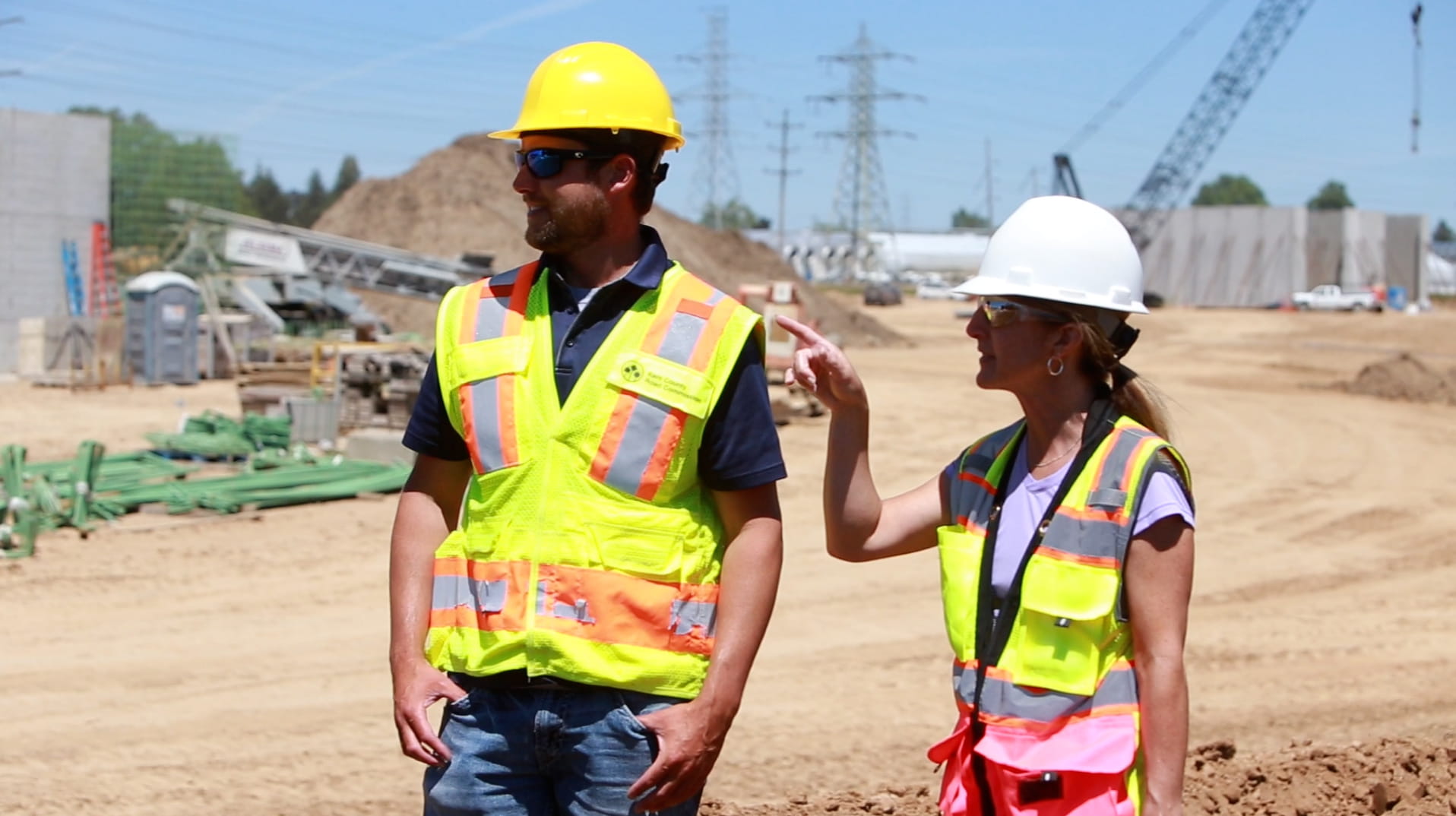 Two industry people standing outside at a worksite, both wearing bright yellow construction vests and hard hats