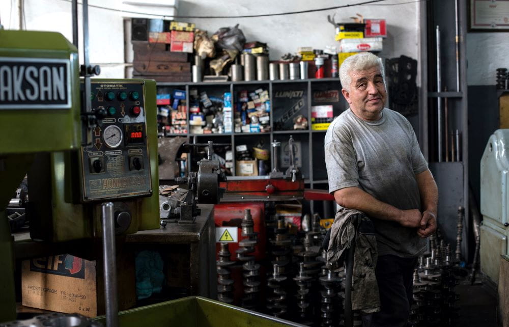 An older man in a grease-stained shirt, leaning against the counter of a mechanic shop, representing local small business