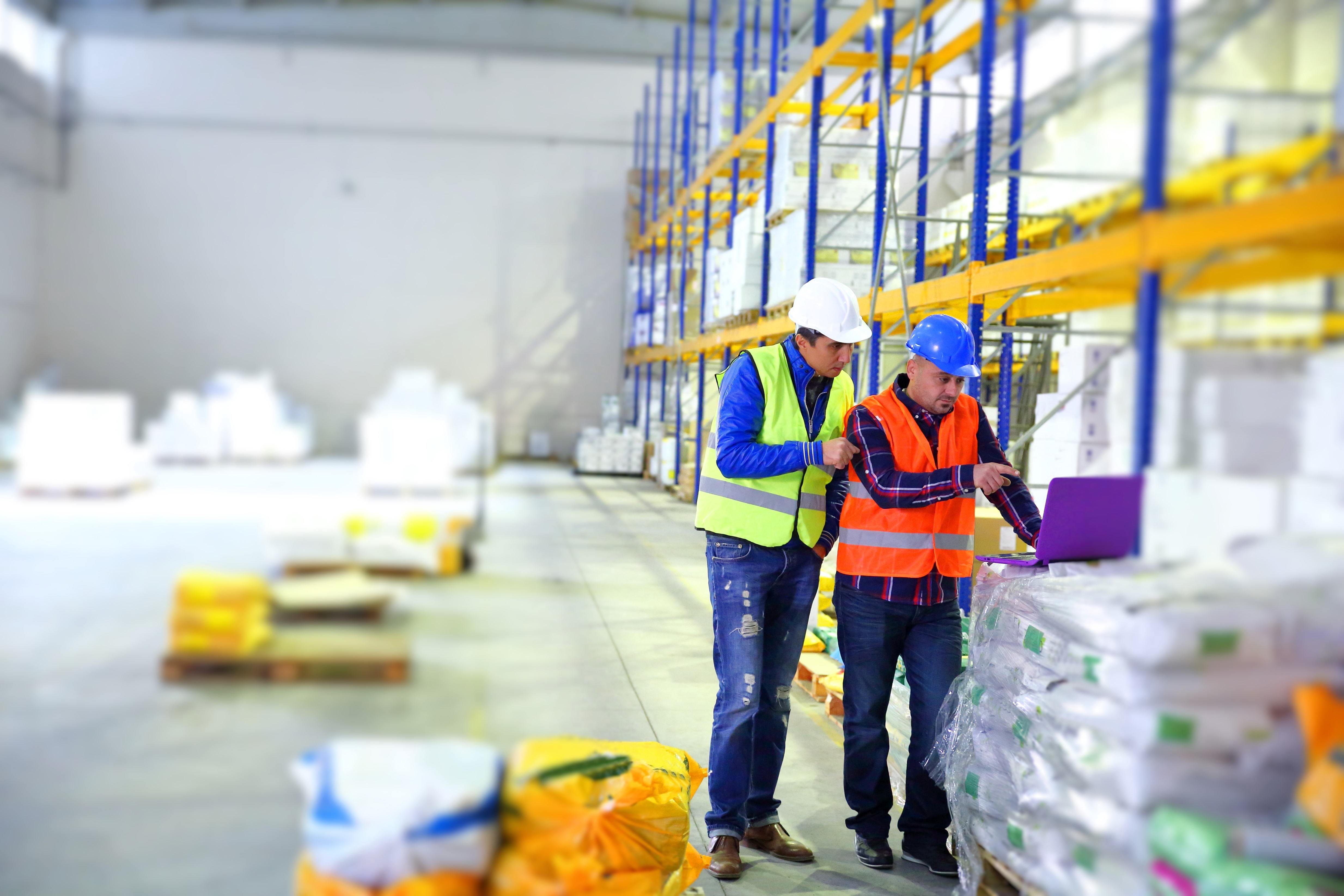 two people in safety vests stand by stored bags of product looking at a laptop