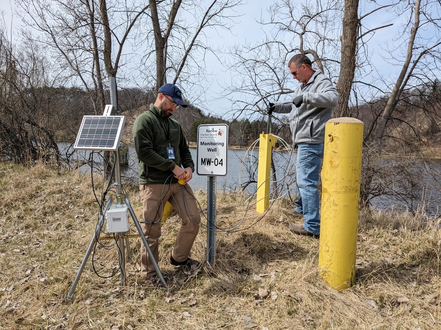 Two people stand on a small hill and install groundwater monitoring equipment on top of a well