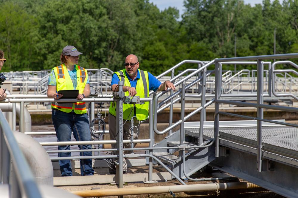 Two EGLE employees stand on a platform above a wastewater treatment tank, one holding a clipboard and the other holding equipment.