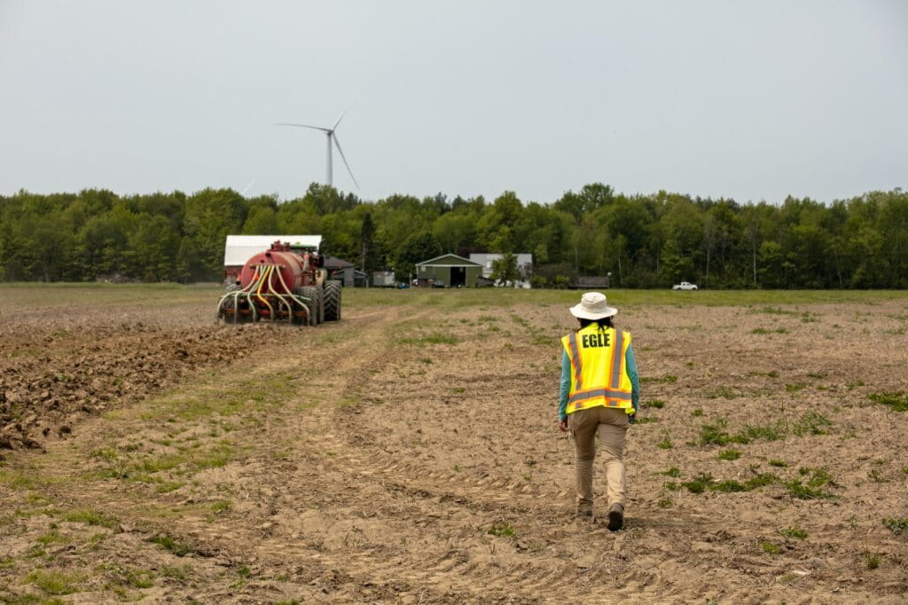 A large machine called a TerraGator driving through a farm field, spraying biosolids directly on the ground through 6 hoses on the back while an EGLE staff person in a yellow vest supervises