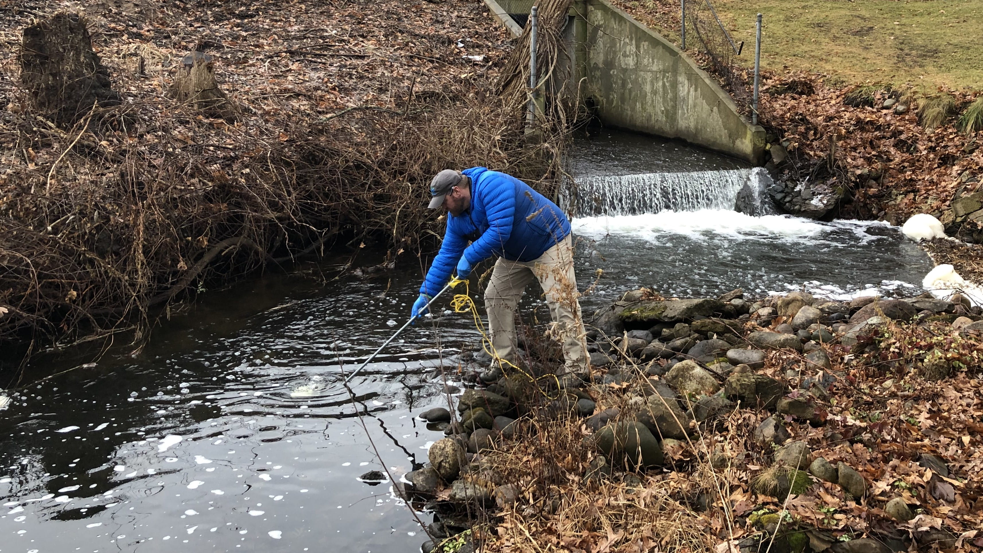 An EGLE contractor stands on the bank of a small culvert, using a long pole (dip pole) to obtain a surface water sample