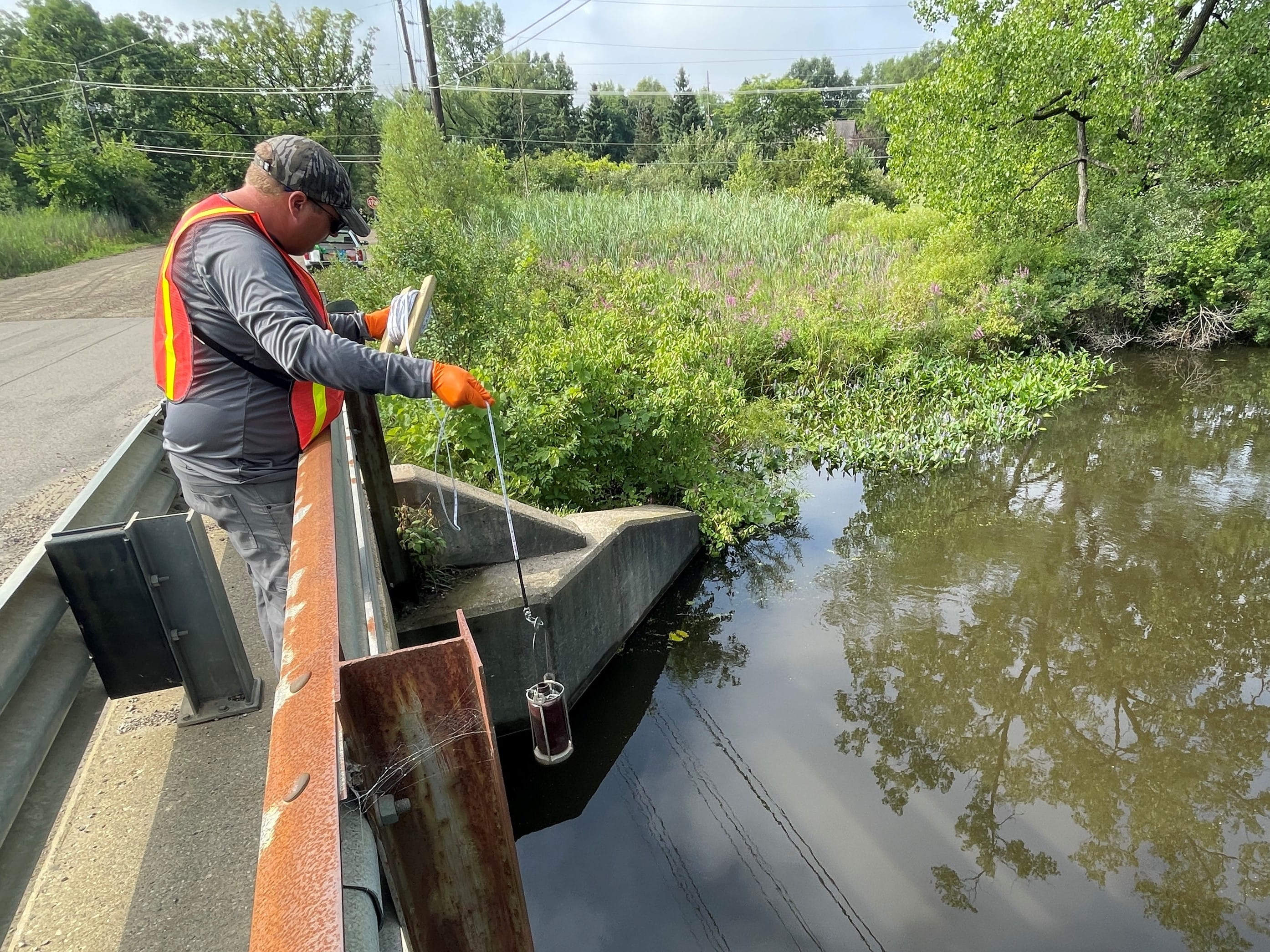 EGLE staff person leaning over an overpass, dipping sampling equipment into the Huron River to collect a water sample due to Tribar Manufacturing release