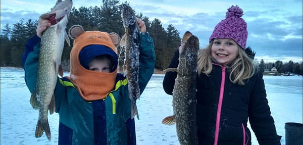 Two children ice fishing, a boy holding two fish and a girl holding one fish