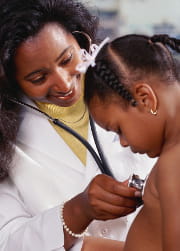 woman doctor in white coat using stethoscope to listen to lungs of young, shirtless girl seated on exam table