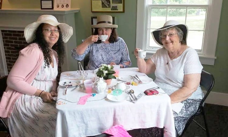 3 women in dresses, hats, talk and sip tea at a linen-covered table with a floral centerpiece. 3 women in dresses, hats, talk and sip tea at a linen-covered table with a floral centerpiece.
