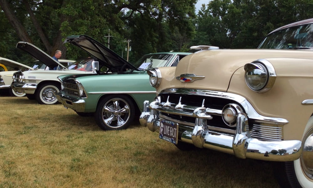 3 classic cars, 2 with hoods up; a man looks at engine of cream-colored mid-20th-century model. 3 classic cars, 2 with hoods up; a man looks at engine of cream-colored mid-20th-century model.