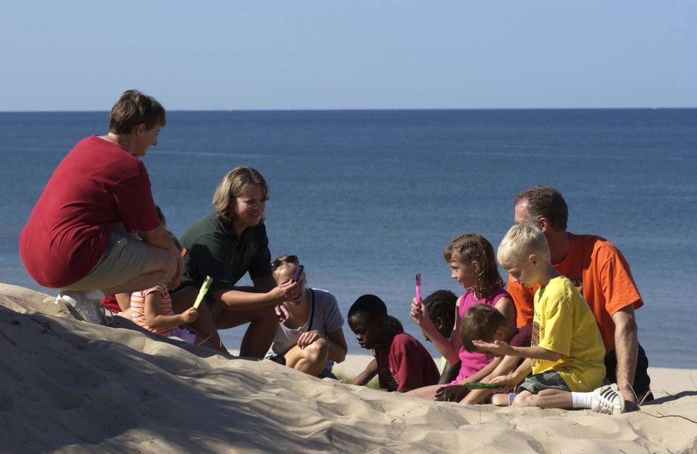 A group of parents and young students on a sand dune above Lake Michigan listen to a DNR educator.
