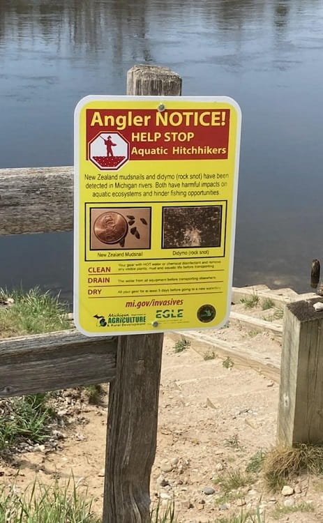 A yellow and red sign on a wood post near a boat launch warning anglers about didymo and New Zealand mudsnails