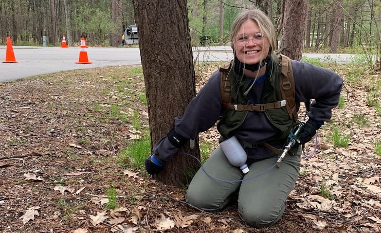 A certified applicator wearing gear kneels near a hemlock tree trunk.
