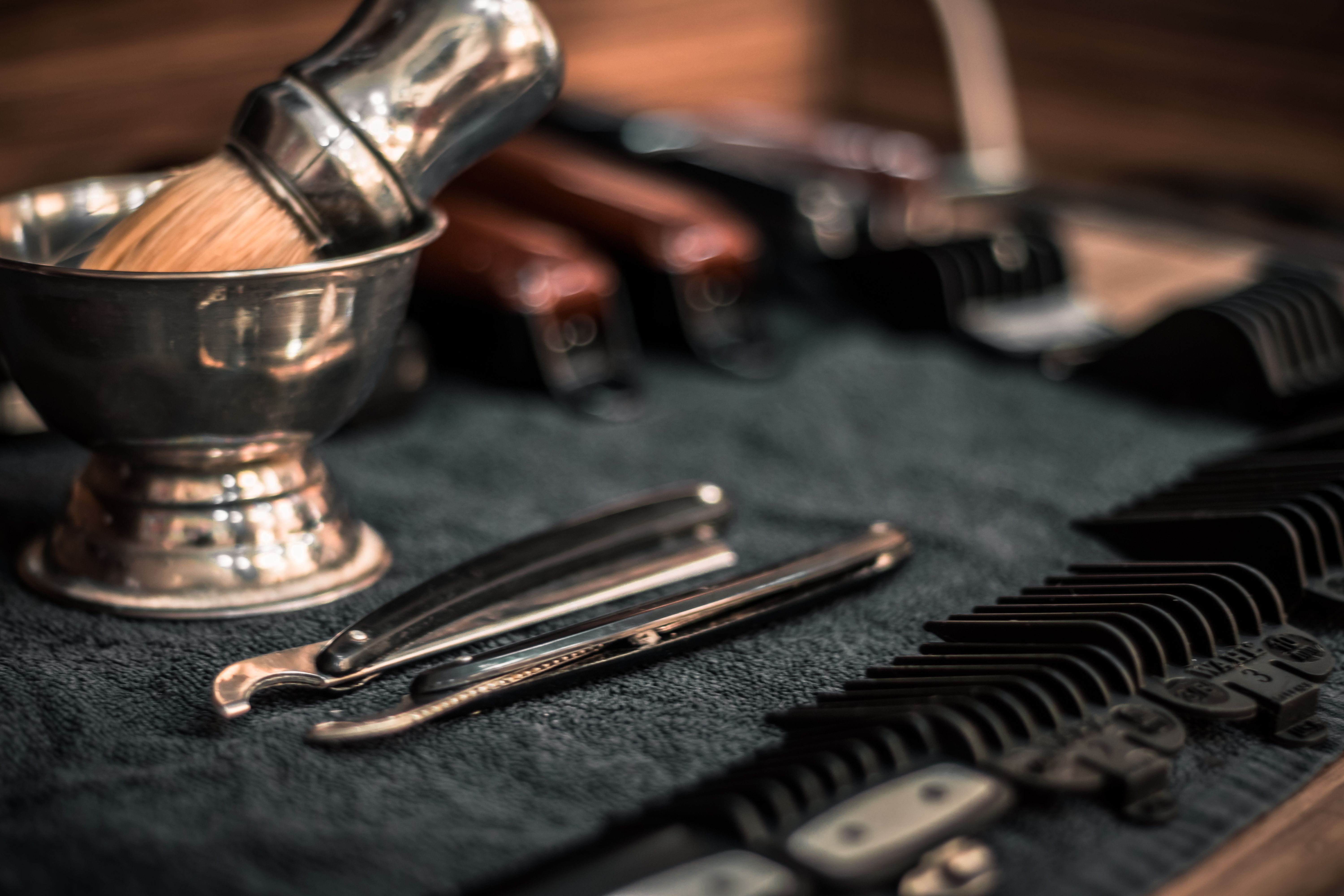 Barber tools such as clippers, guard combs, straight razors and a shaving brush with bowl laid out on a towel