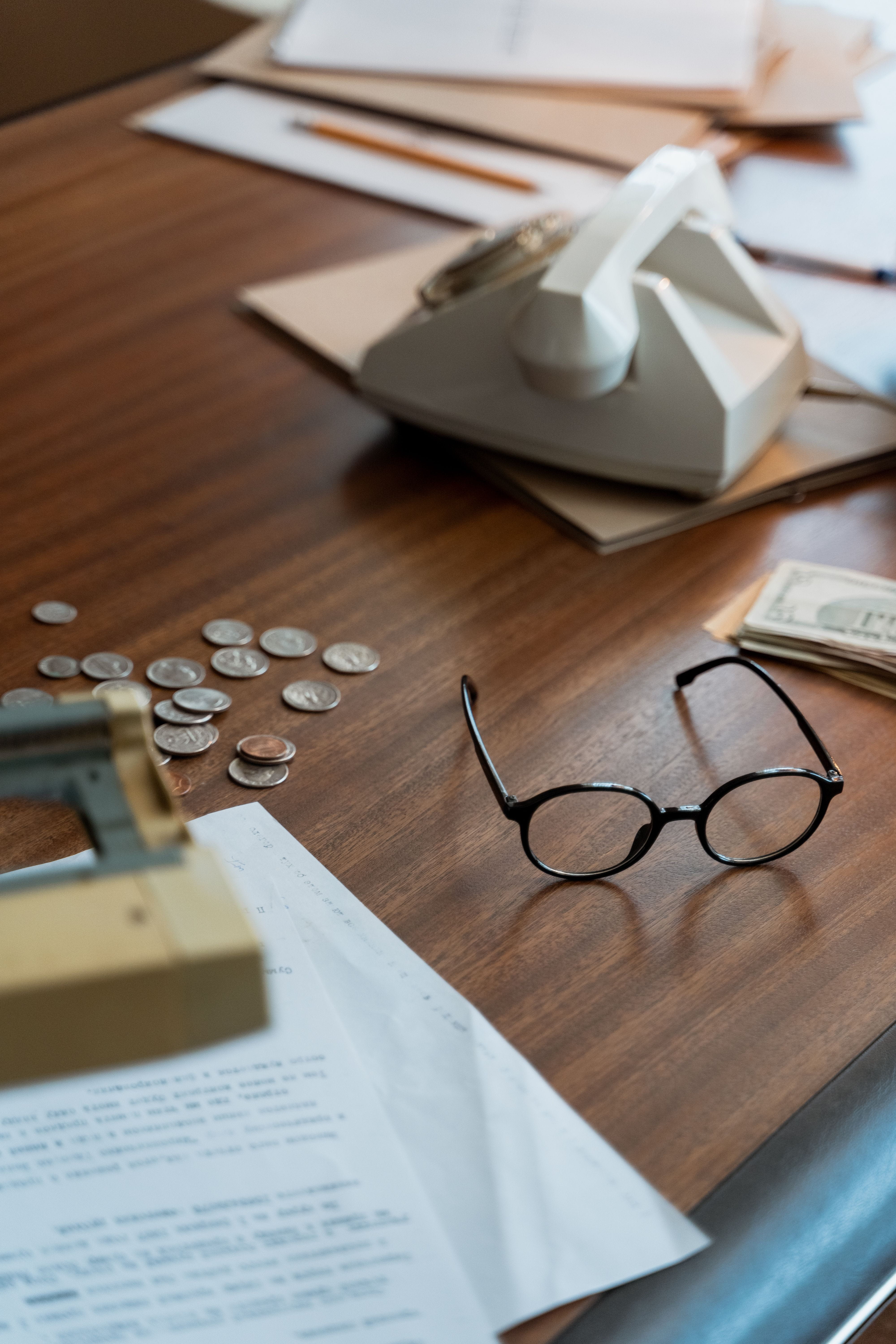 Desk with folders, paperwork, desk phone, money and a pair of eye glasses