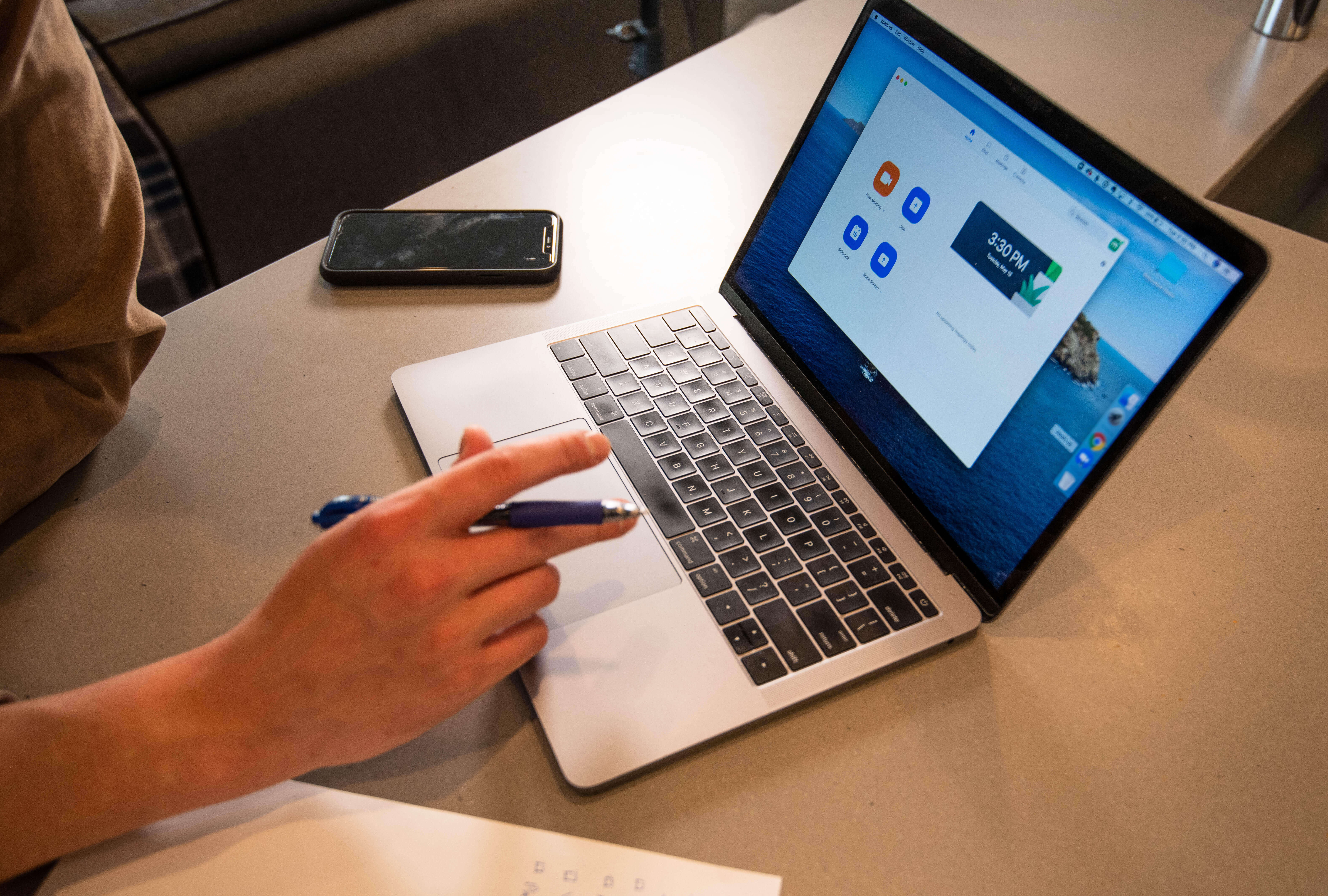 Hand holding a pen while using a laptop with a video meeting app open; a phone and notes sit on the table.