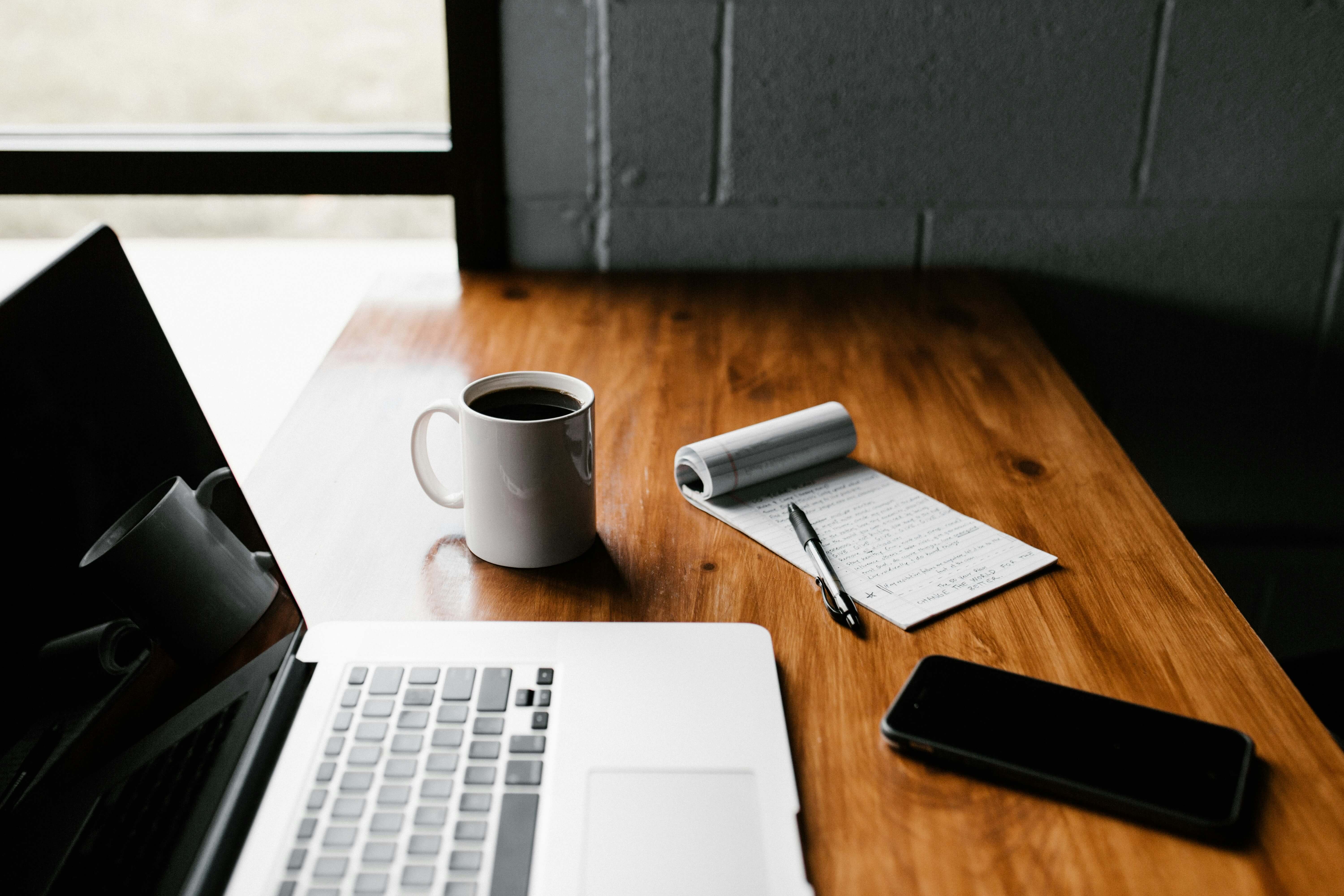 A Laptop, phone, writing pad, pen, and a cup of coffee sitting on a desk.