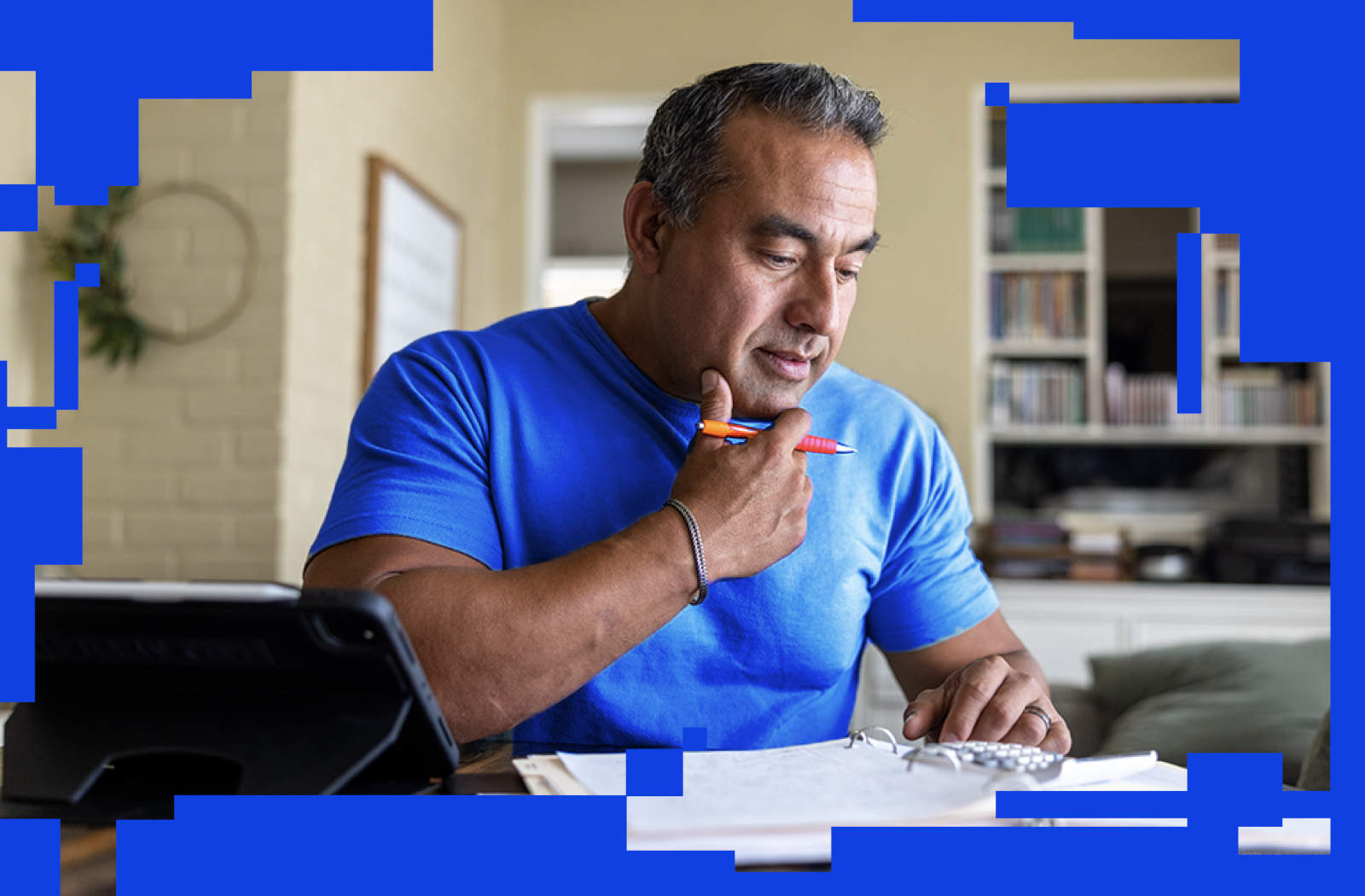 An individual sits at a desk in a home office, holding a pencil while looking at documents on the table. The desk contains papers, a calculator, and a device positioned in the foreground. Bookshelves and home décor items are visible in the background. The image is framed with a blue geometric border made up of rectangular cutouts.