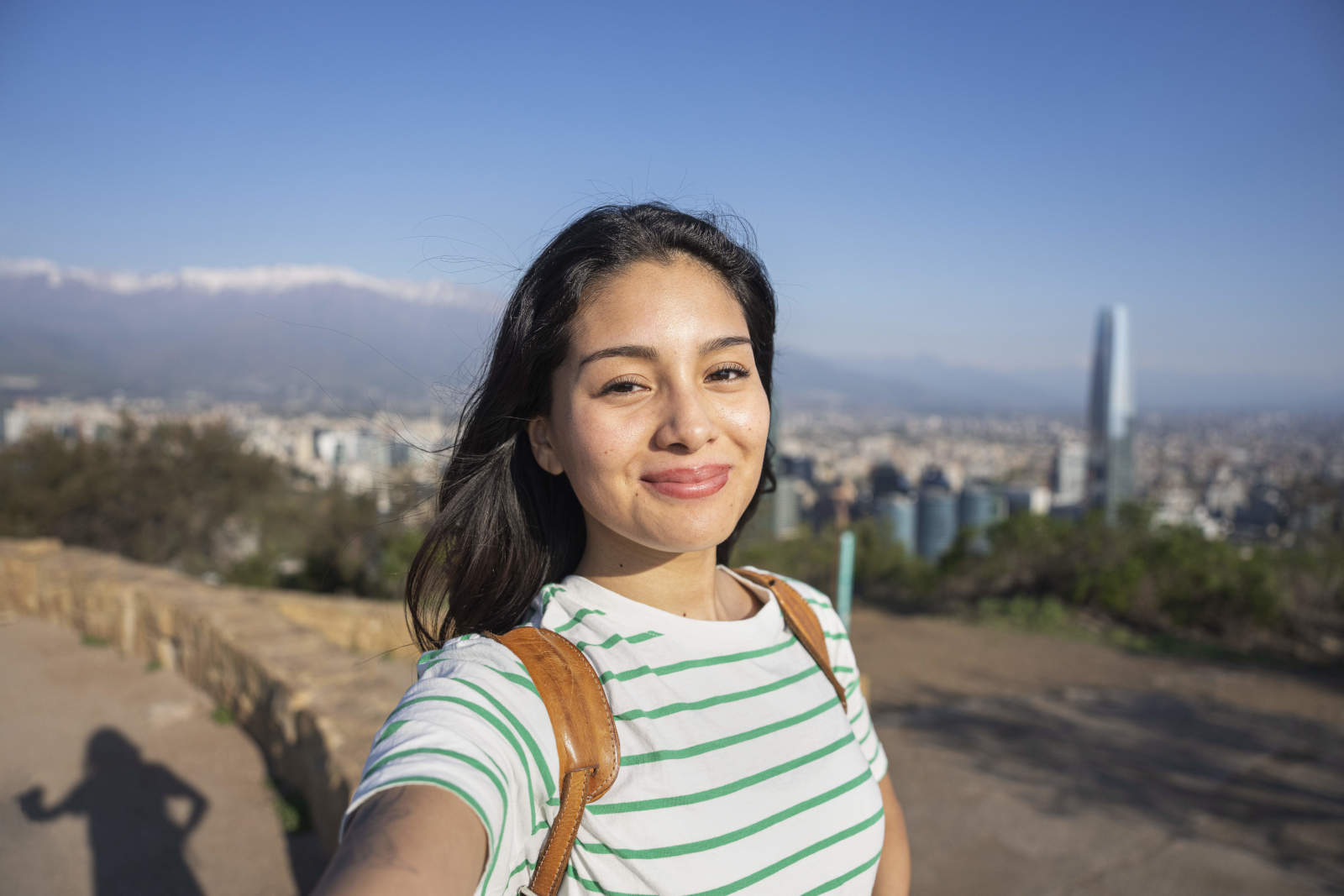 The image shows an individual taking a selfie outdoors on a sunny day. The person is wearing a striped shirt and a backpack strap is visible over one shoulder. In the background, there is a cityscape with multiple buildings, a tall tower, and distant mountains partially covered with snow. The scene includes a stone wall, a dirt pathway, and greenery along the hillside under a clear blue sky.
