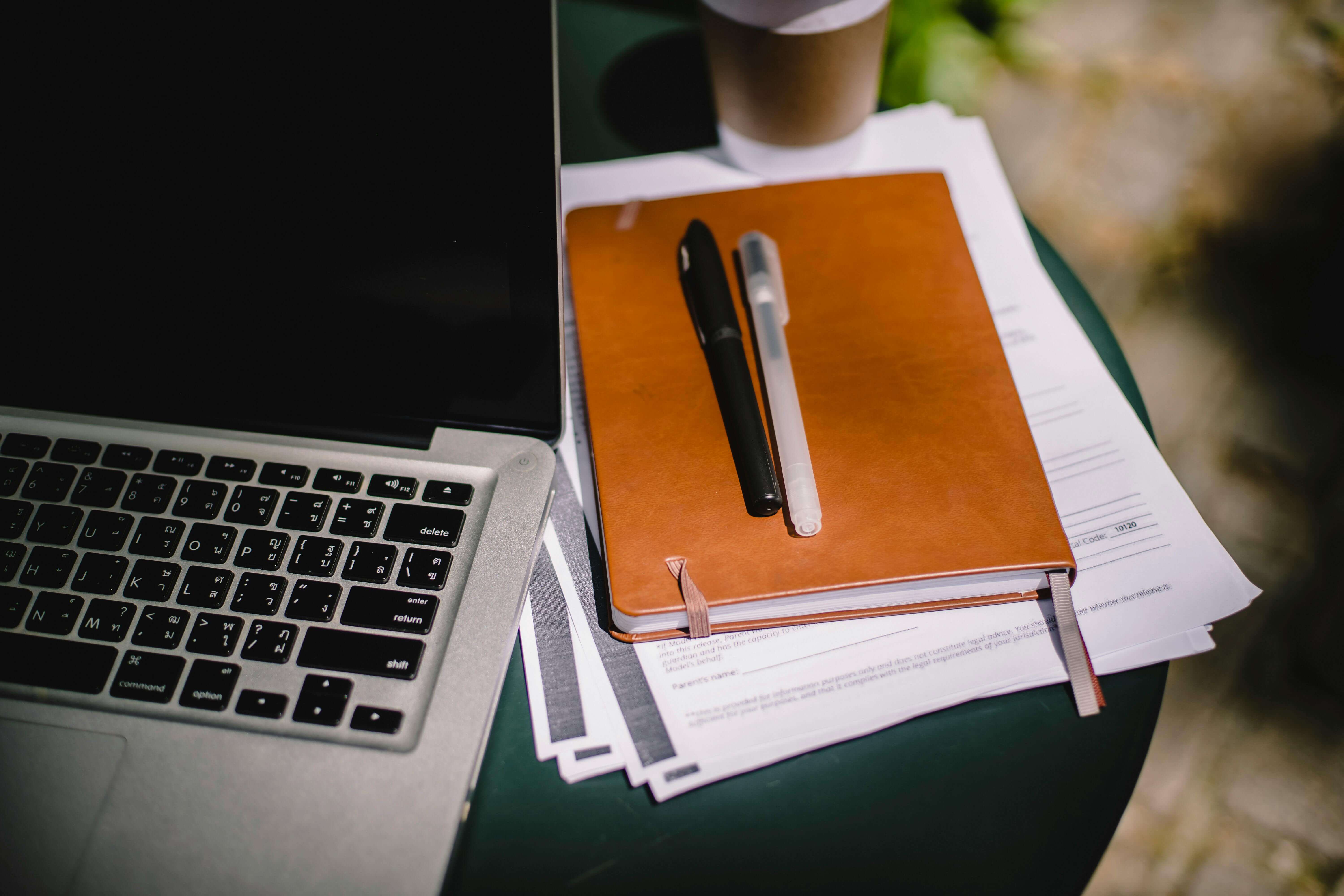 Two pens stacked on top of a notebook and multiple documents sitting next to a laptop