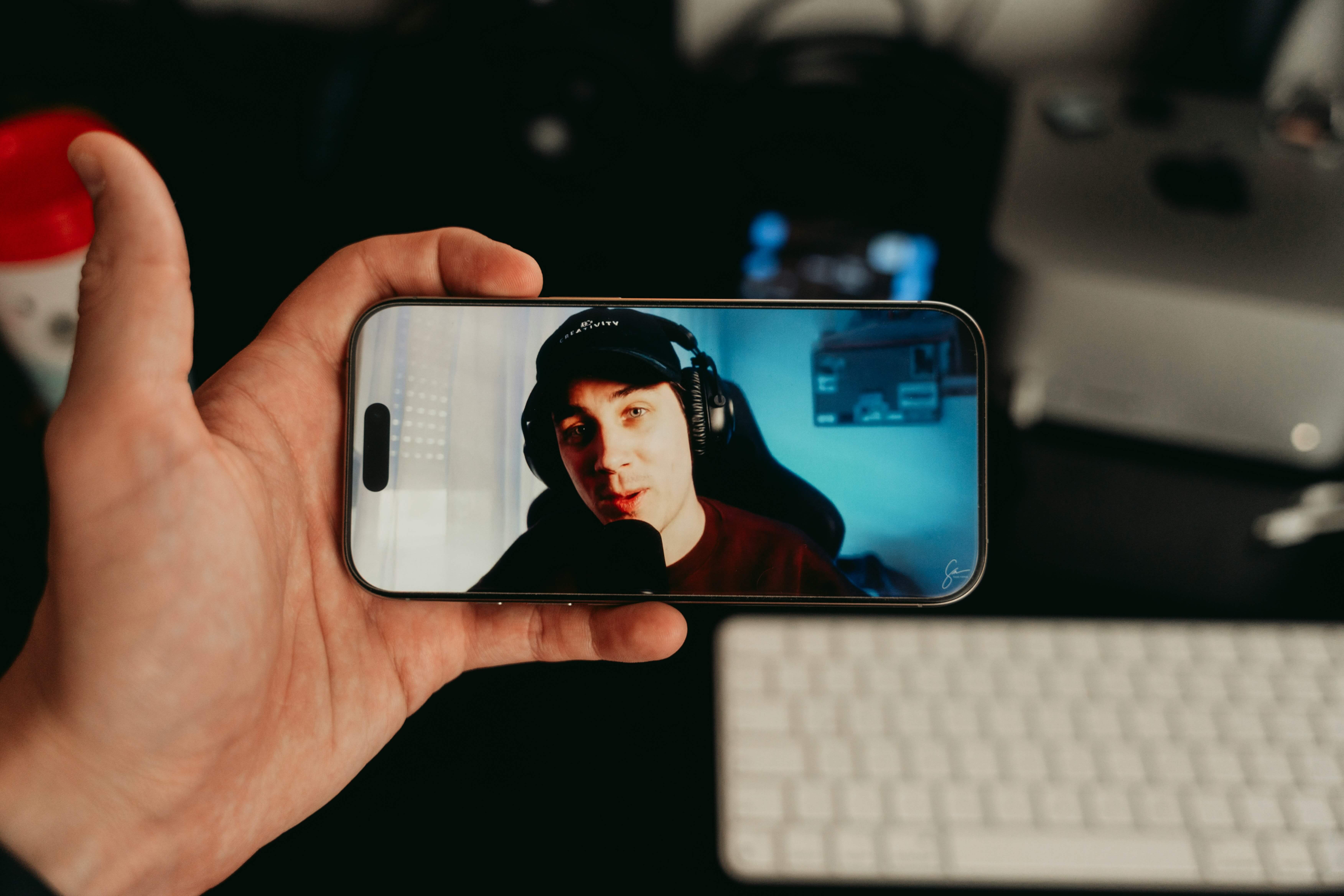 A hand is holding a smartphone displaying a person wearing large over‑ear headphones and a dark top, seated in front of a gaming-style setup with a blue-lit screen in the background. The scene is indoors on a desk with a white keyboard, computer monitor, and other tech accessories visible behind the phone.