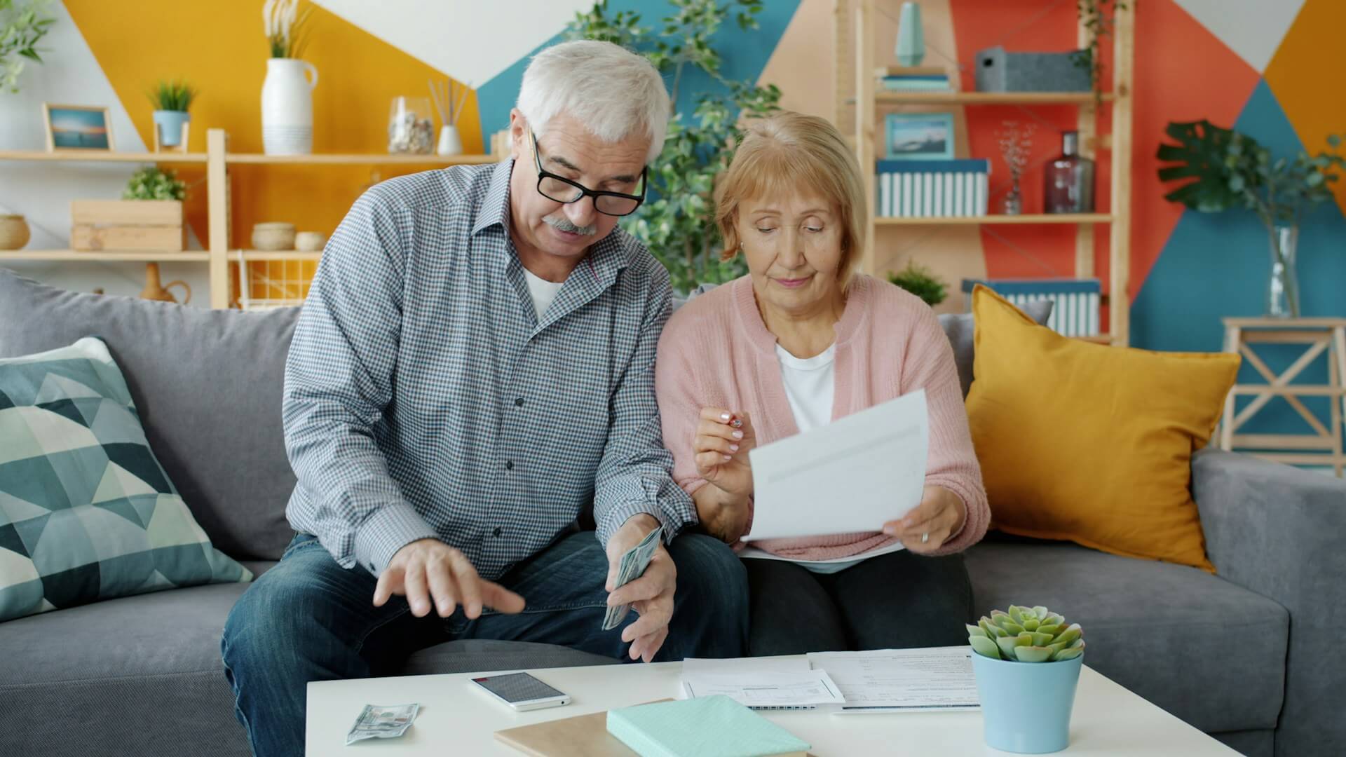 Two older adults sit on a couch reviewing financial documents together. One holds cash and gestures toward a smartphone on the table, while the other looks at a sheet of paper. Papers, a notebook, and a small plant sit on the coffee table in front of them.