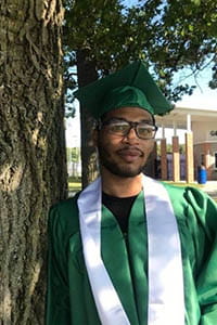 A person wearing a green graduation hat and glasses standing next to a tree
                                            