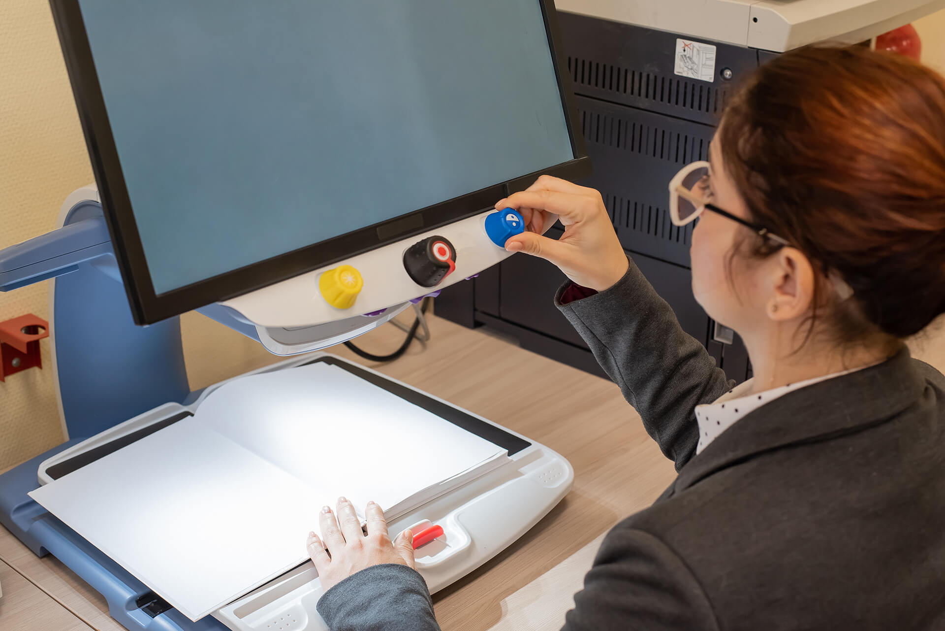 A person adjusts a blue control knob on a large electronic magnifier positioned above an open book on a desktop workstation.