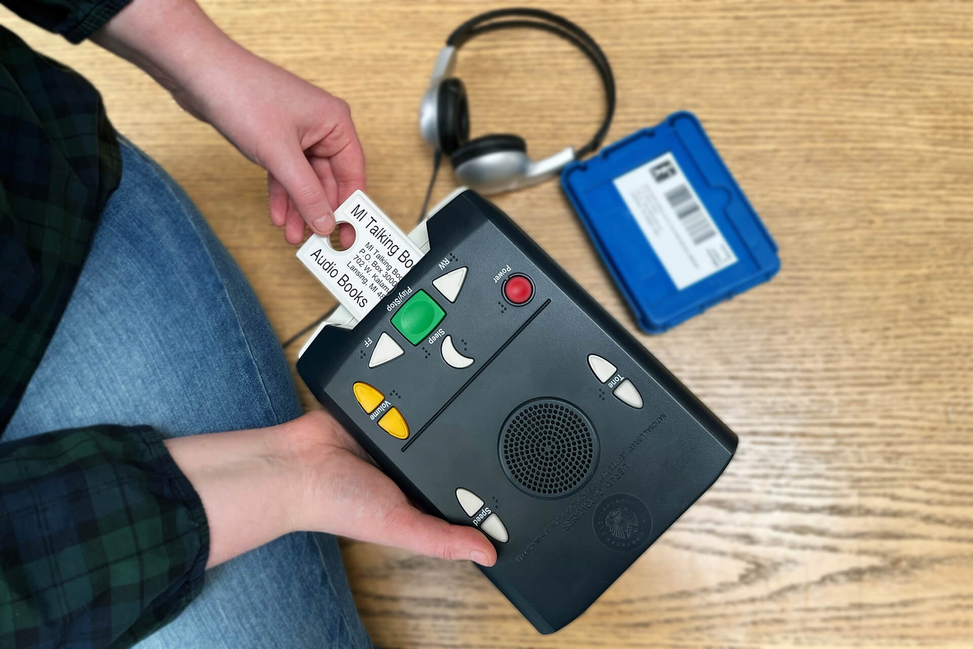A person inserts an audio book cartridge labeled “MI Talking Books” into a digital talking‑book player, with headphones and a blue storage case nearby.
