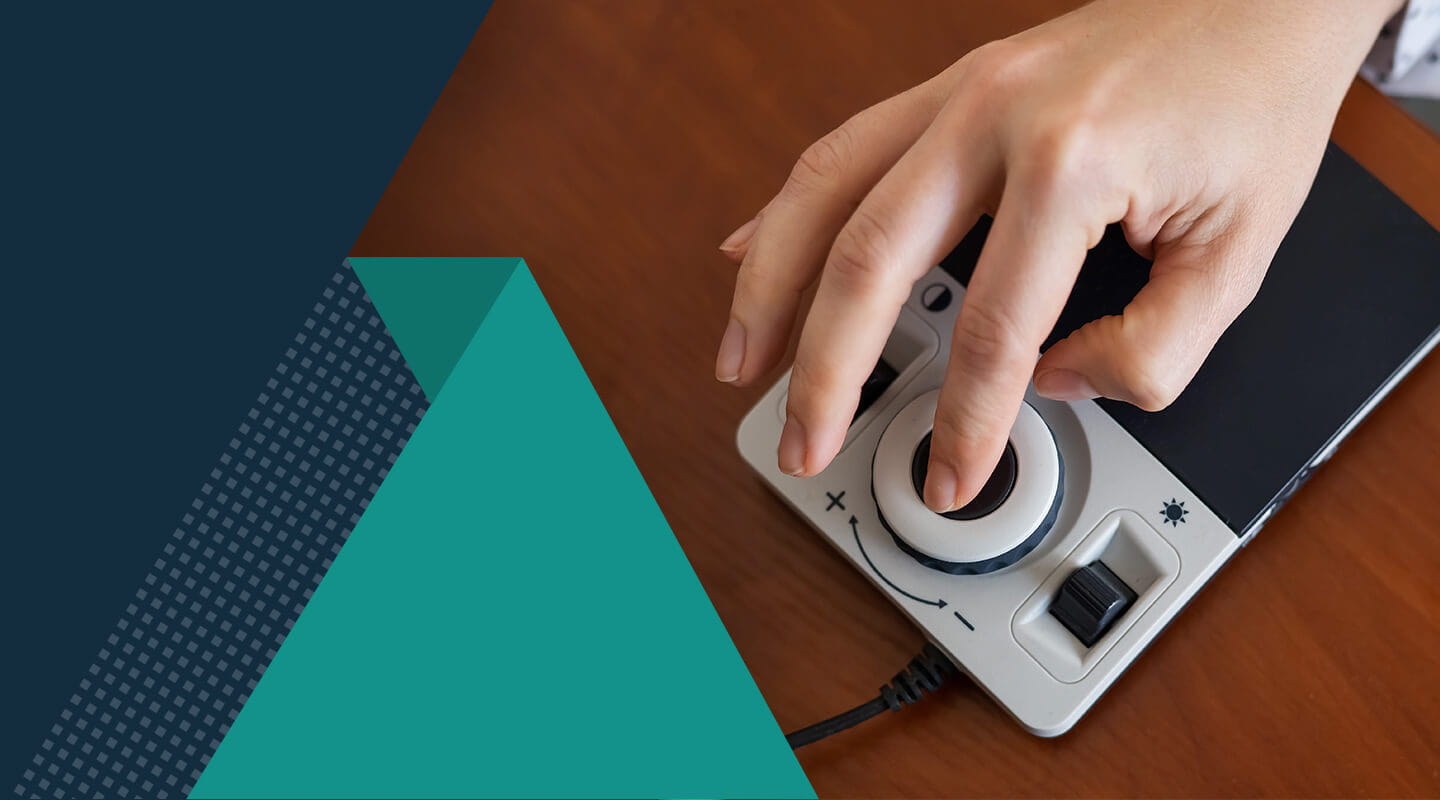 A hand operates an adaptive electronic device featuring a large circular control dial and additional buttons, positioned on a wooden surface. Geometric teal and dark blue shapes overlay part of the image.