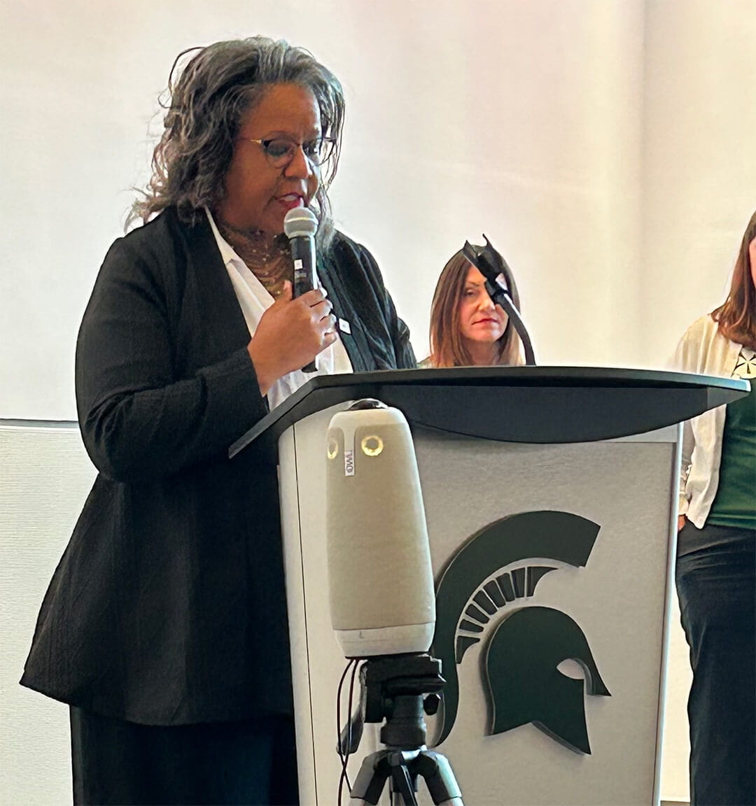 Sigrid Adams, bureau director for Michigan Rehabilitation Services, speaks into a handheld microphone at a podium with a Spartan logo, with two women standing behind her at the 2025 Abilities, Opportunities and Future of Work Conference.